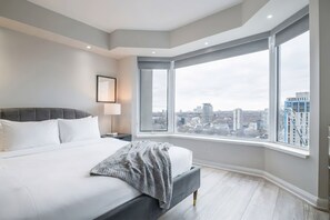 Un dormitorio moderno con una cama grande, una mesita de noche, una lámpara y vistas a la ciudad a través de amplios ventanales.
