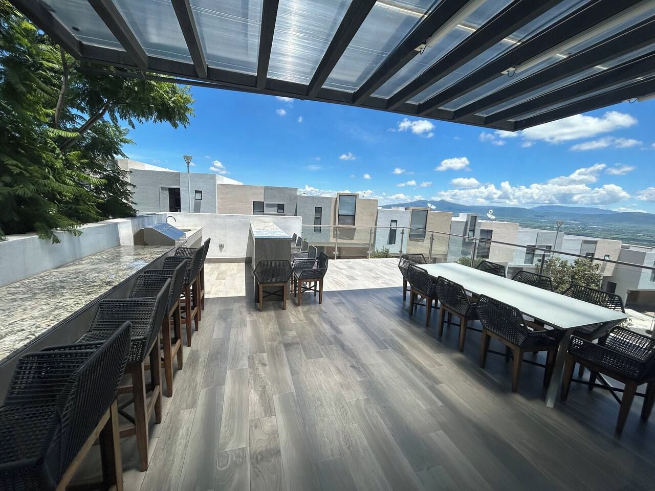 Une terrasse extérieure avec une longue table et des chaises, une pergola au-dessus, et une vue sur les bâtiments et les montagnes.
