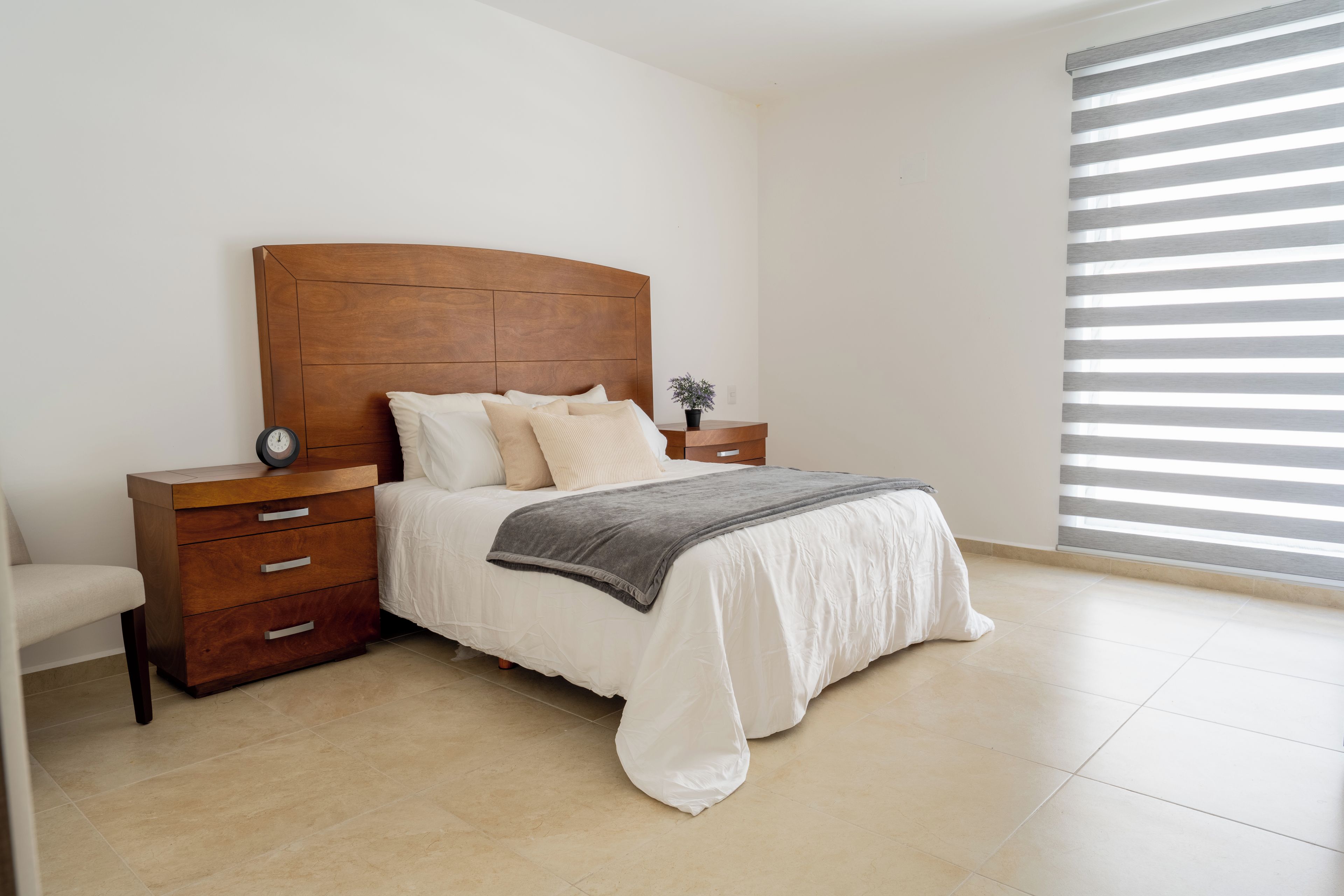 Une chambre à coucher avec une tête de lit en bois, un lit avec une literie blanche, une table de chevet avec une horloge et une fenêtre avec des stores horizontaux.