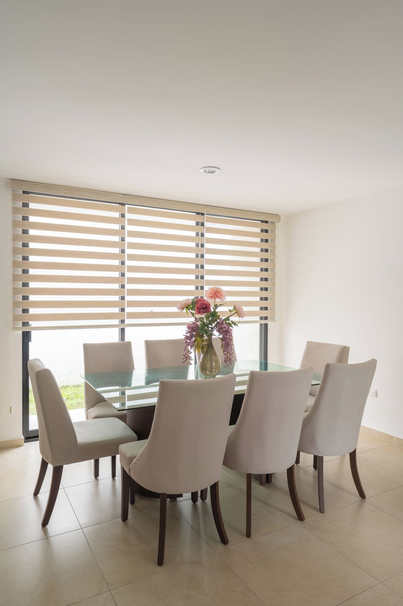 A dining area with a glass table surrounded by chairs, a vase with flowers, and wooden blinds on the windows.