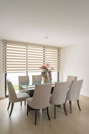 Un coin repas avec une table en verre entourée de chaises, un vase de fleurs et des stores en bois aux fenêtres.