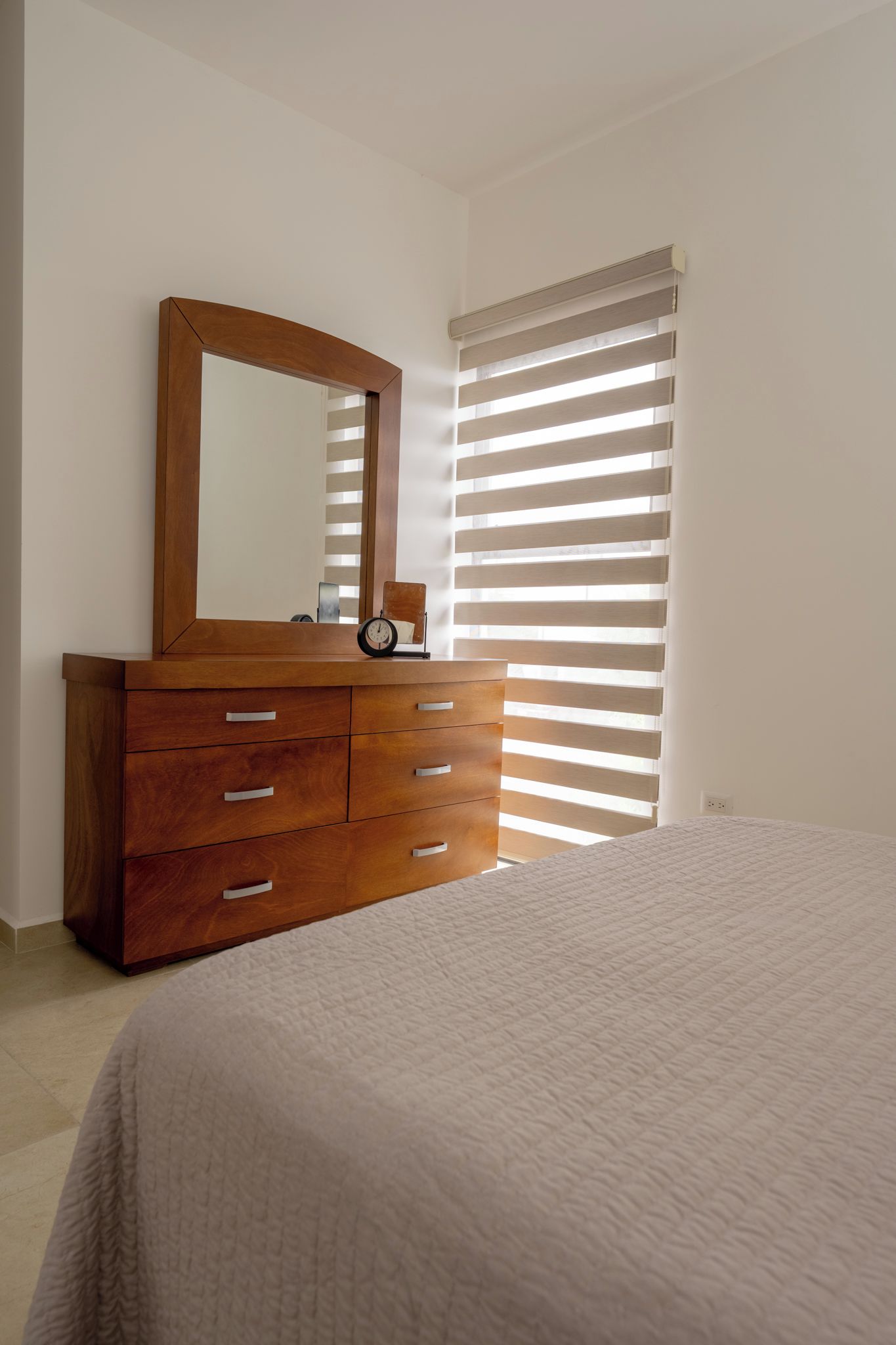Une chambre à coucher avec une commode en bois, un lit recouvert d’une courtepointe blanche, un miroir et une fenêtre avec des stores horizontaux.