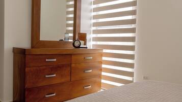 Une chambre à coucher avec une commode en bois, un lit recouvert d’une courtepointe blanche, un miroir et une fenêtre avec des stores horizontaux.