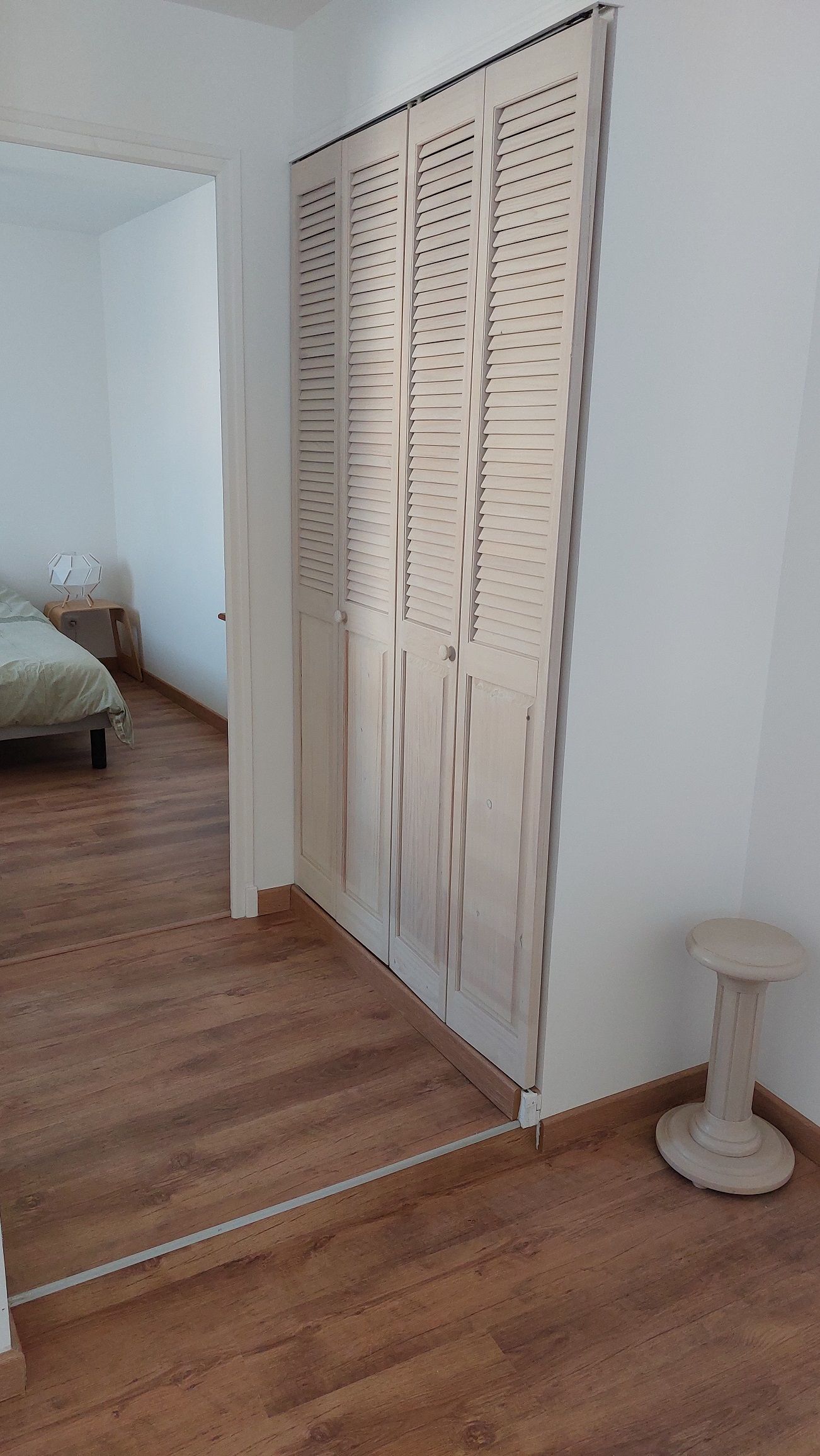 Une chambre avec un plancher en bois, un lit, une armoire à portes à persiennes et une petite table ronde.