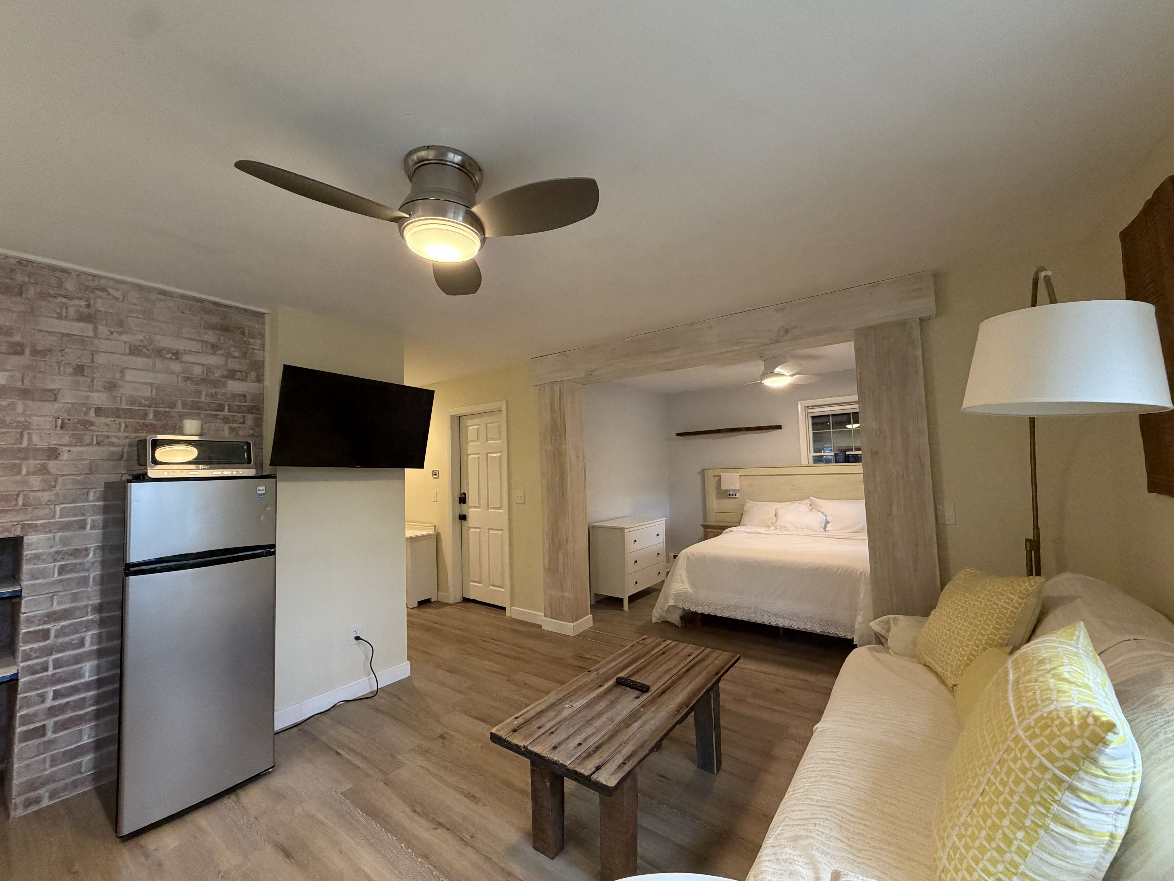 Une chambre d’hôtel compacte comprenant un coin cuisine, un lit, une table de chevet, un ventilateur de plafond et une lampe.