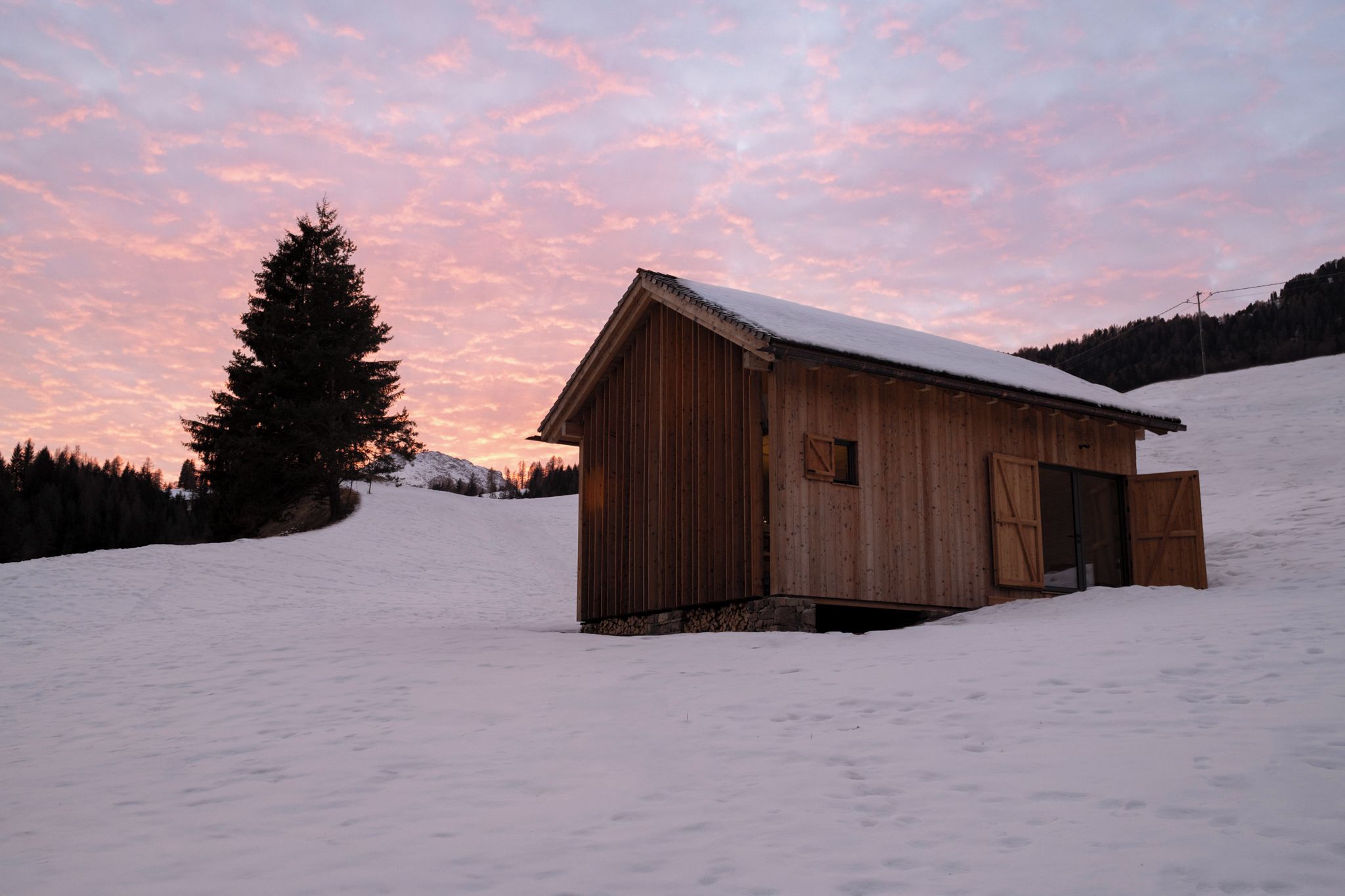 A wooden cabin with a snow-covered roof, situated on a snow-covered slope with a backdrop of a sunset sky.