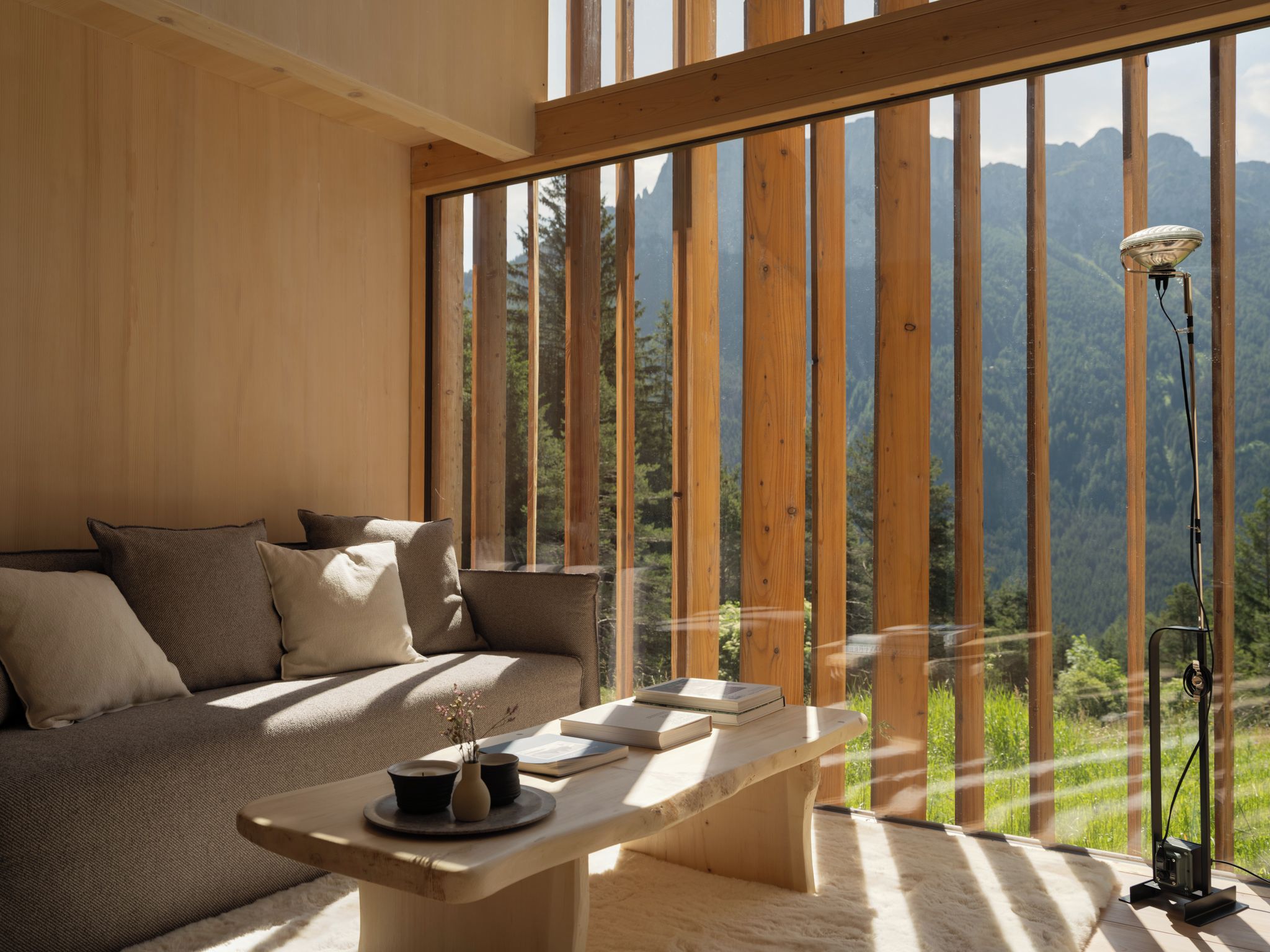 A room with a wooden floor, a sofa, a table with books and candles, and a view of mountains through large windows.