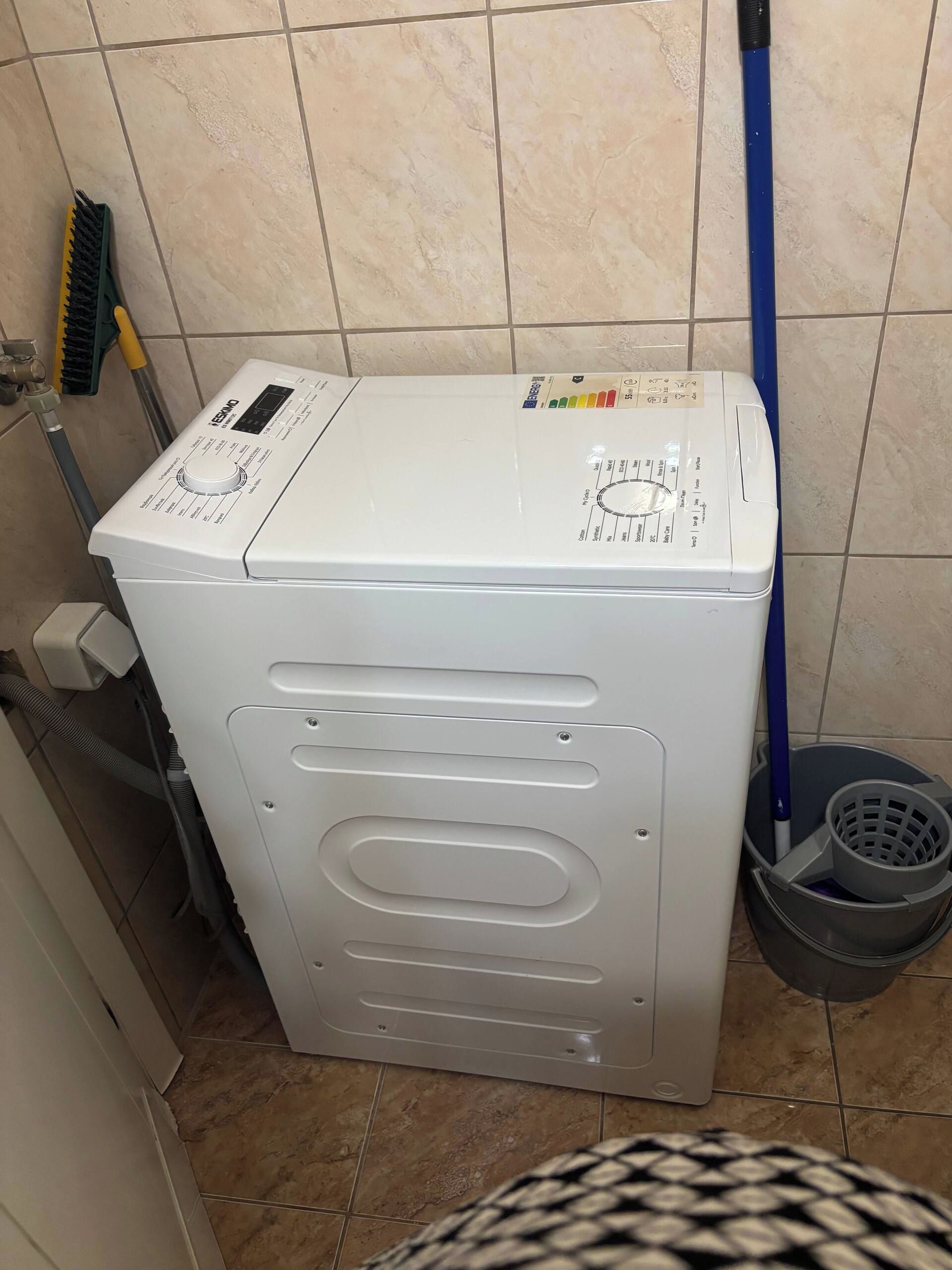 Una màquina de rentar blanca en un bany amb parets de rajola i una fregona adossada a la paret.