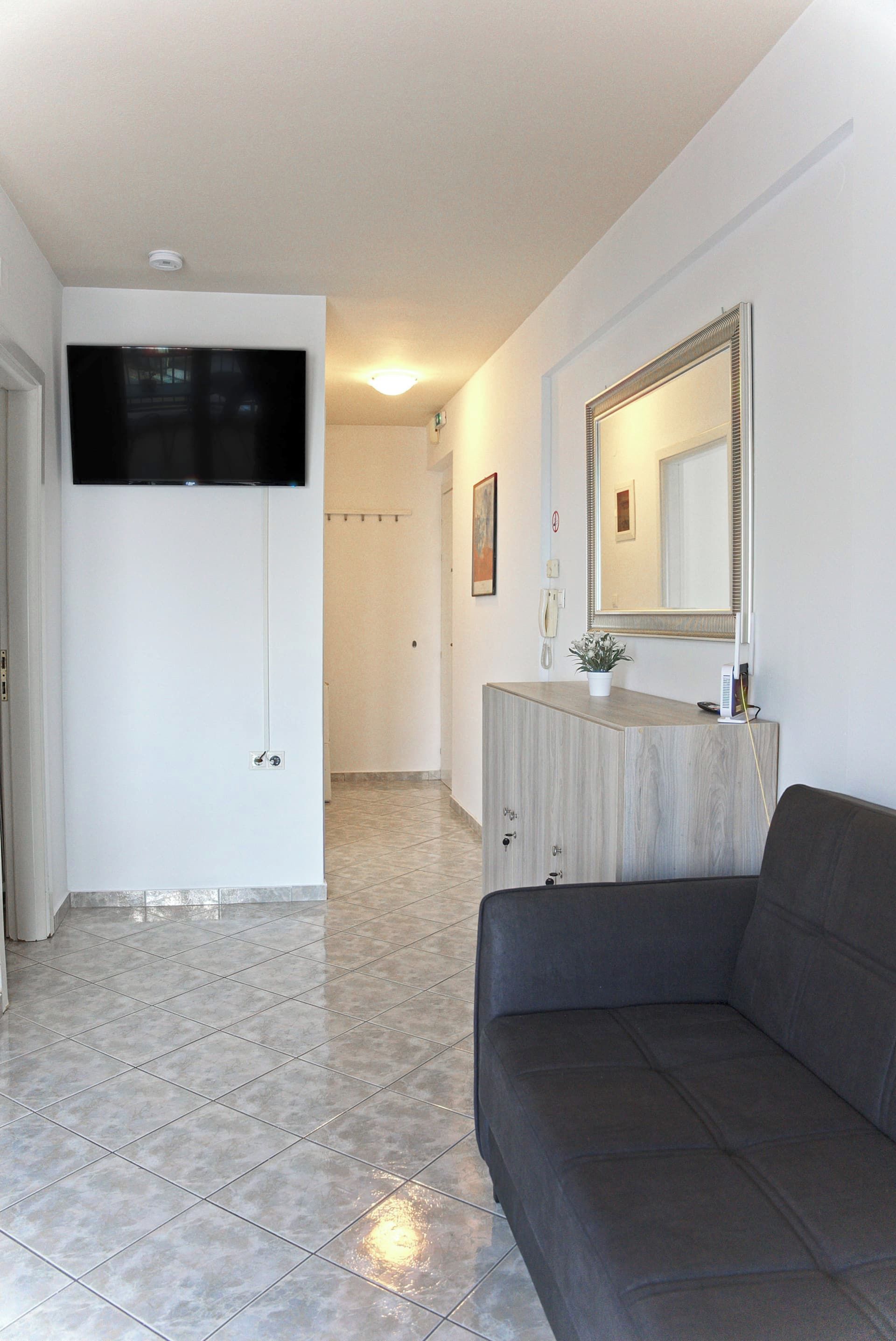 Ein Hotelzimmer mit einem an der Wand befestigten Flachbildfernseher, einem Holzschrank mit Spiegel, einem grauen Sofa und einem gefliesten Boden.