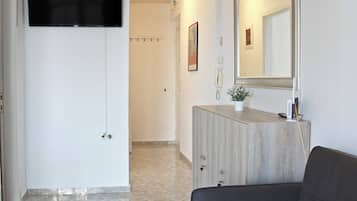 Ein Hotelzimmer mit einem an der Wand befestigten Flachbildfernseher, einem Holzschrank mit Spiegel, einem grauen Sofa und einem gefliesten Boden.