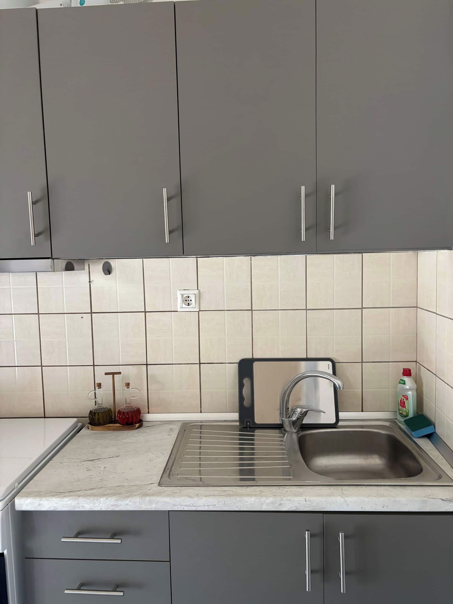 Cuina amb armaris grisos, un rentamiqueses d'acer inoxidable i un mur revestit de rajoles.