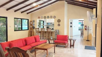 A spacious living area with a high ceiling, wooden furniture, and a kitchen in the background.
