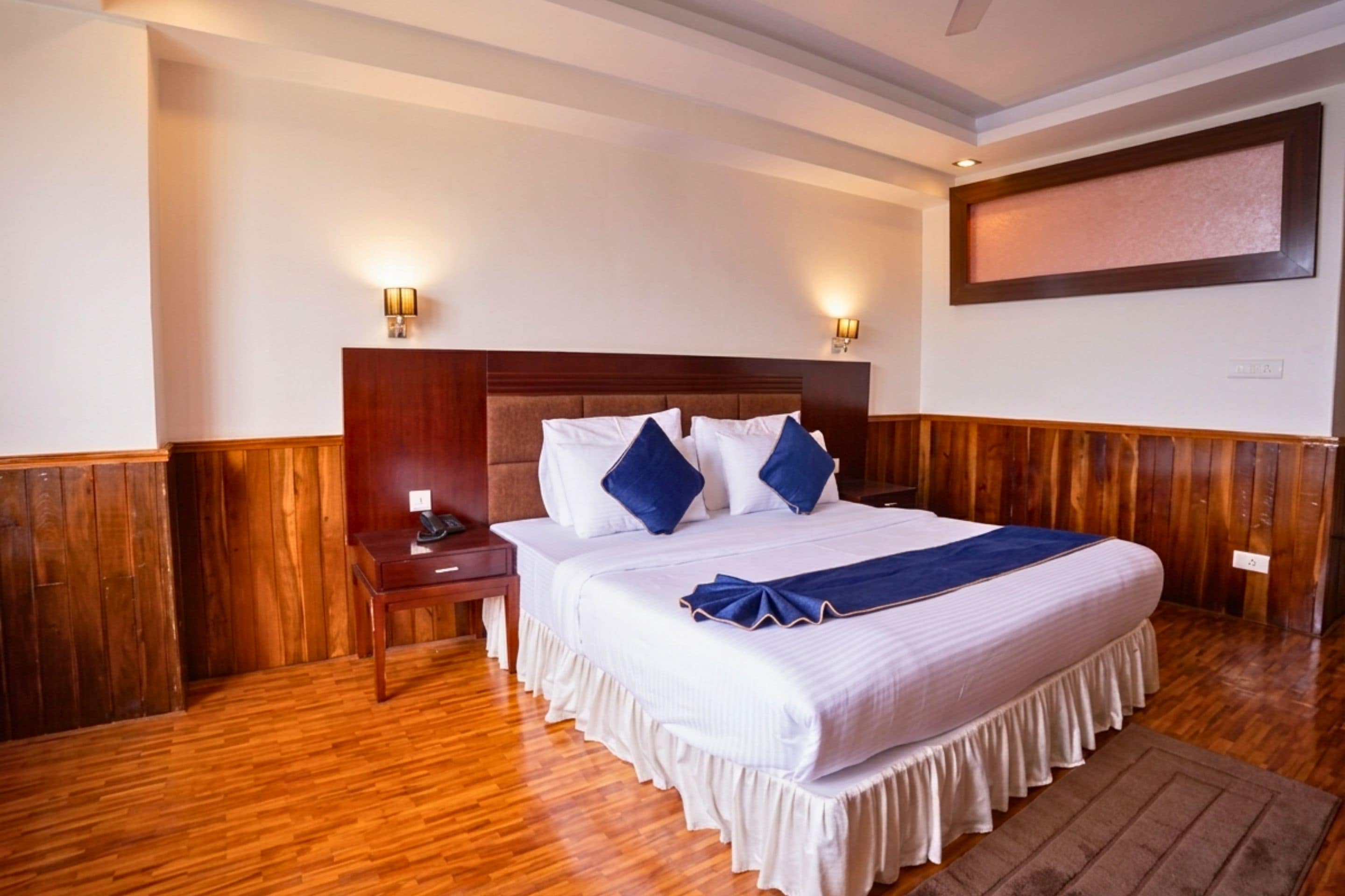 A hotel room with a wooden floor, a large bed with white bedding and blue pillows, two bedside tables with lamps, and a large framed picture on the wall.