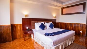 A hotel room with a wooden floor, a large bed with white bedding and blue pillows, two bedside tables with lamps, and a large framed picture on the wall.