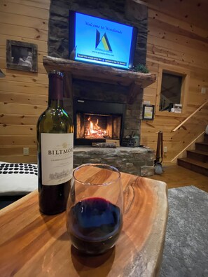 Un interno in legno con camino, un portatile, una bottiglia di vino Biltmore e un bicchiere di vino rosso su un vassoio di legno.
