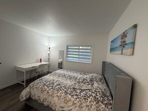 Een slaapkamer met een bed, een bureau, een stoel, een spiegel, een raam met zonwering en een schilderij met strandthema aan de muur.