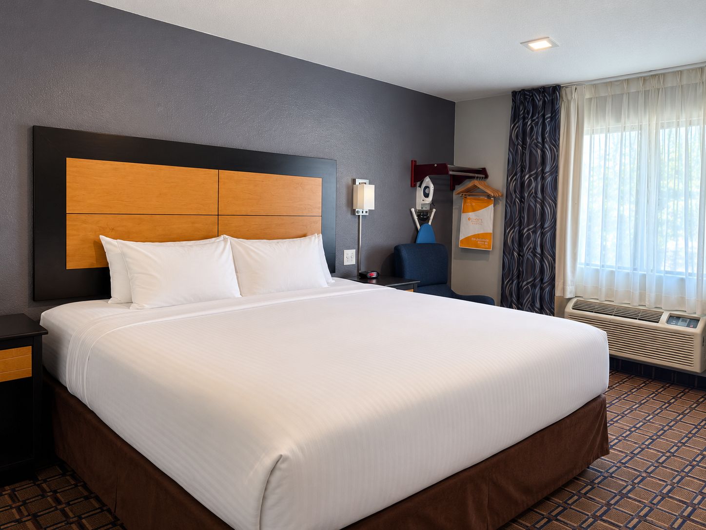 Une chambre d’hôtel avec un grand lit, une table de chevet, un bureau, une chaise, une fenêtre avec des rideaux et un radiateur fixé au mur.