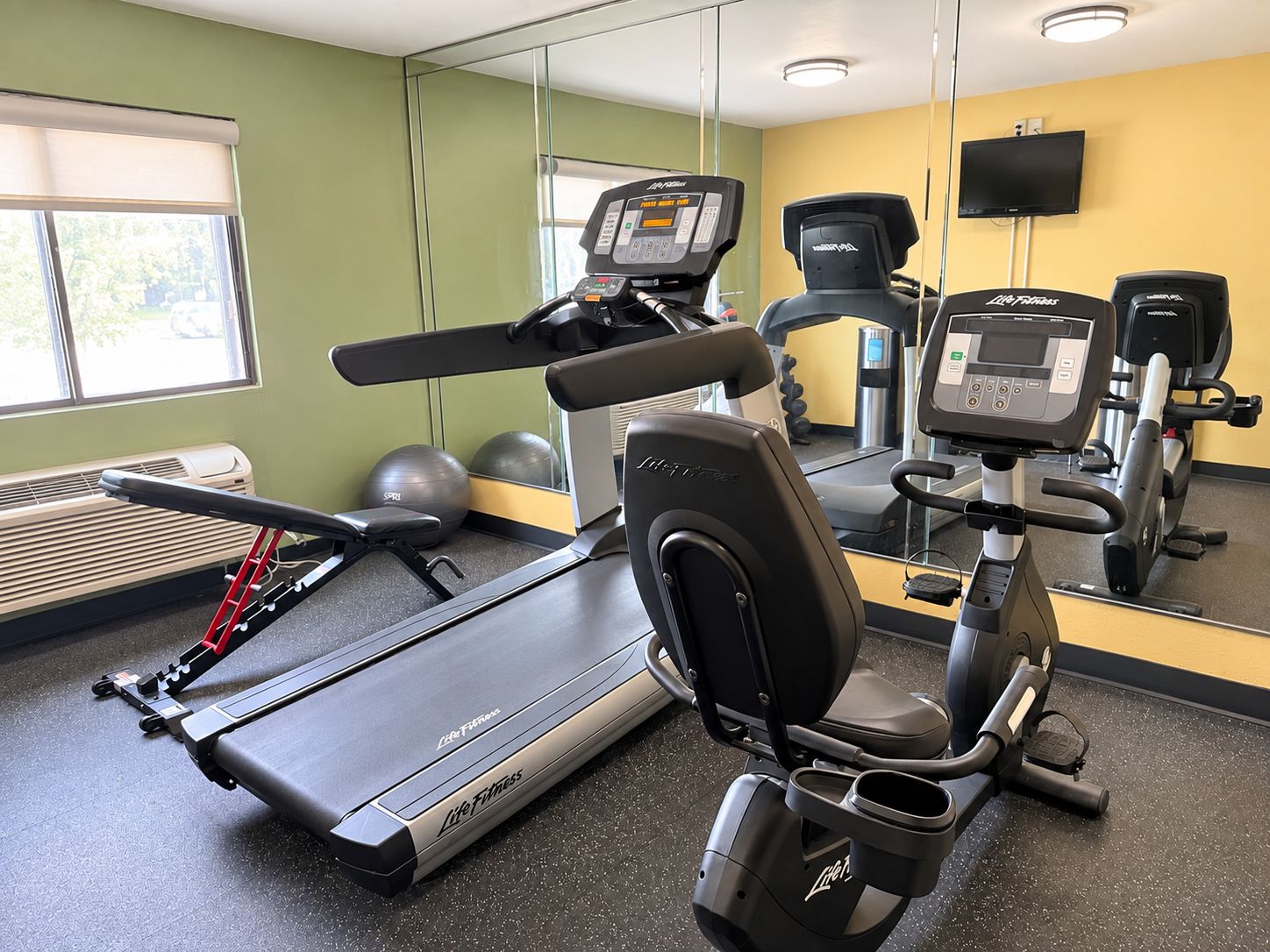 Ein Fitnessraum mit Cardiogeräten, darunter ein Laufband und ein Crosstrainer, einem Spiegel und einem Fernseher an der Wand.