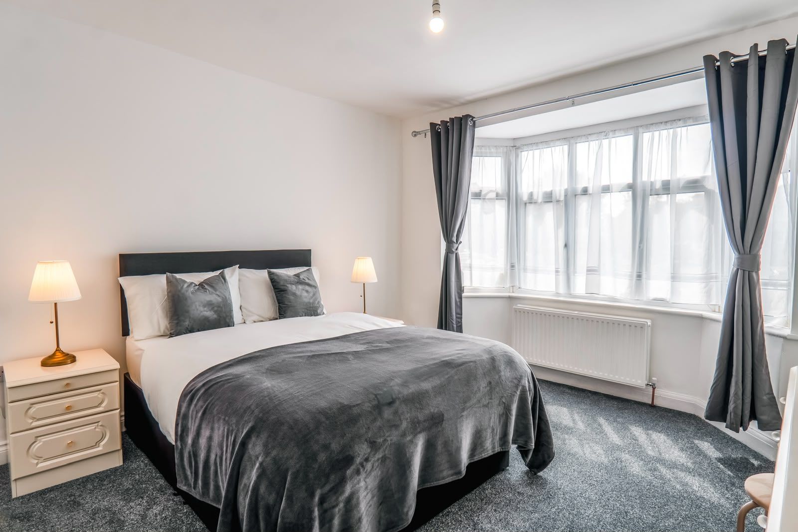Une chambre d’hôtel avec un lit, des lampes de chevet, une fenêtre avec des rideaux, un radiateur et un sol recouvert de moquette.