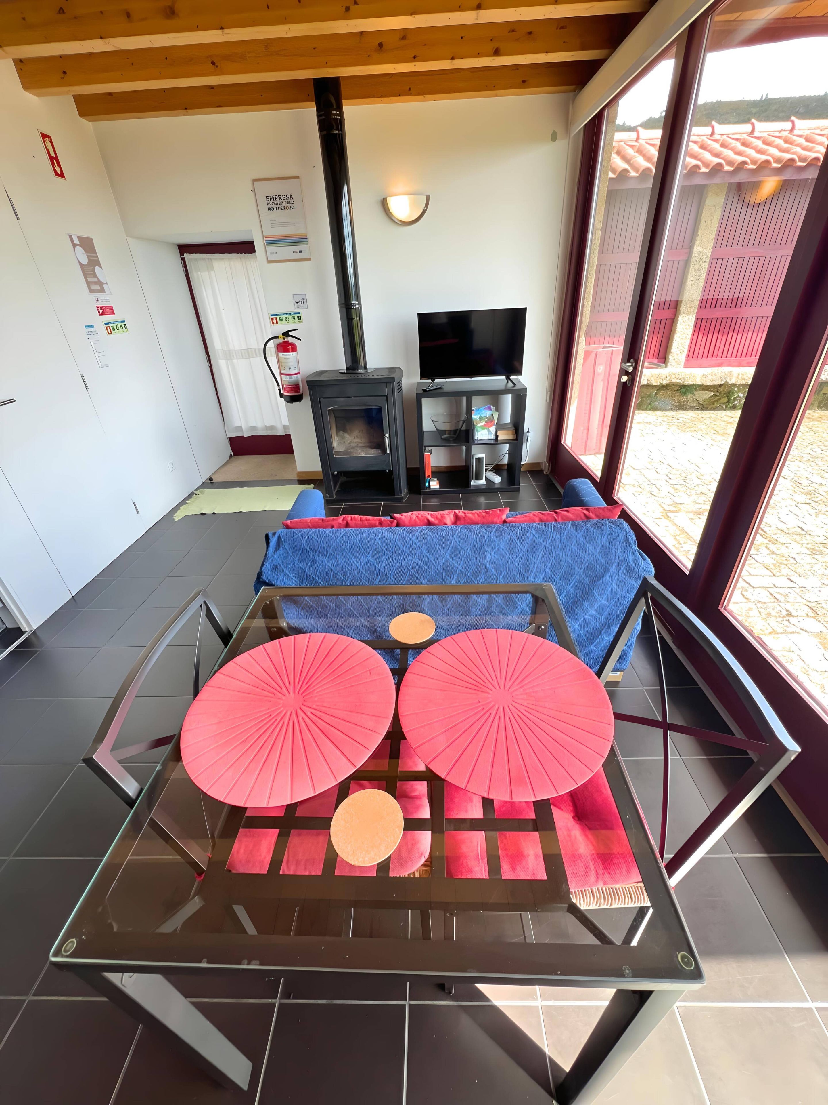 Una habitación con chimenea, televisor y una mesa de vidrio con cojines rojos.