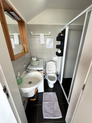 Un baño moderno con lavamanos, inodoro y espejo blancos. Hay un perchero con una toalla y una pequeña estantería con artículos de tocador. El piso está cubierto de baldosas, y hay un felpudo frente al inodoro.