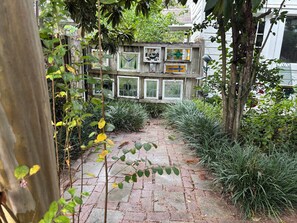 A brick pathway leading to a wooden fence with multiple windows, surrounded by greenery and trees. - Eclectic 2-bedroom house with delightful outside spaces in walkable Montrose (Houston)