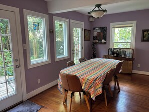 A dining area with a table covered in a striped tablecloth, surrounded by chairs, and large windows with views of greenery. - Eclectic 2-bedroom house with delightful outside spaces in walkable Montrose (Houston)