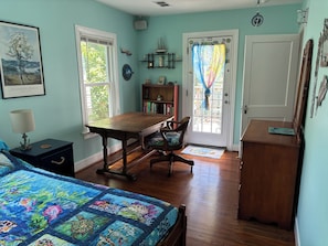 A room with a wooden desk, a chair, a bed with a colorful quilt, a bookshelf, and a door with a curtain. - Eclectic 2-bedroom house with delightful outside spaces in walkable Montrose (Houston)