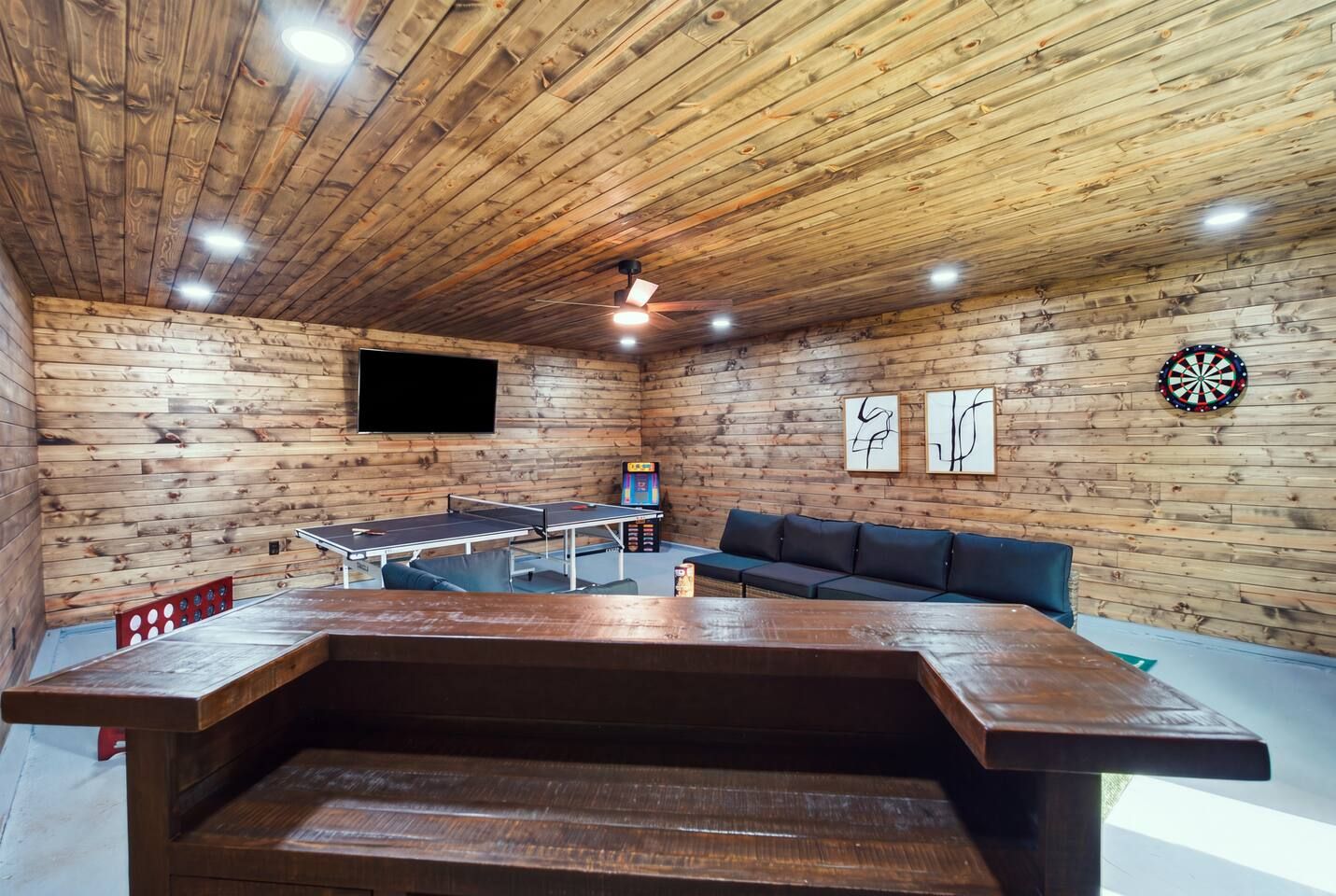 A room with a ping pong table, a sofa, a dartboard, and a flat-screen TV on a wooden wall.