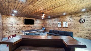A room with a ping pong table, a sofa, a dartboard, and a flat-screen TV on a wooden wall.