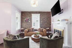 A living room with a brick wall, a fireplace, a television mounted on the wall, and a sofa with multiple cushions. - Hot Tub + 2 Kings | Near KY Horse Park | 3BR (Georgetown)