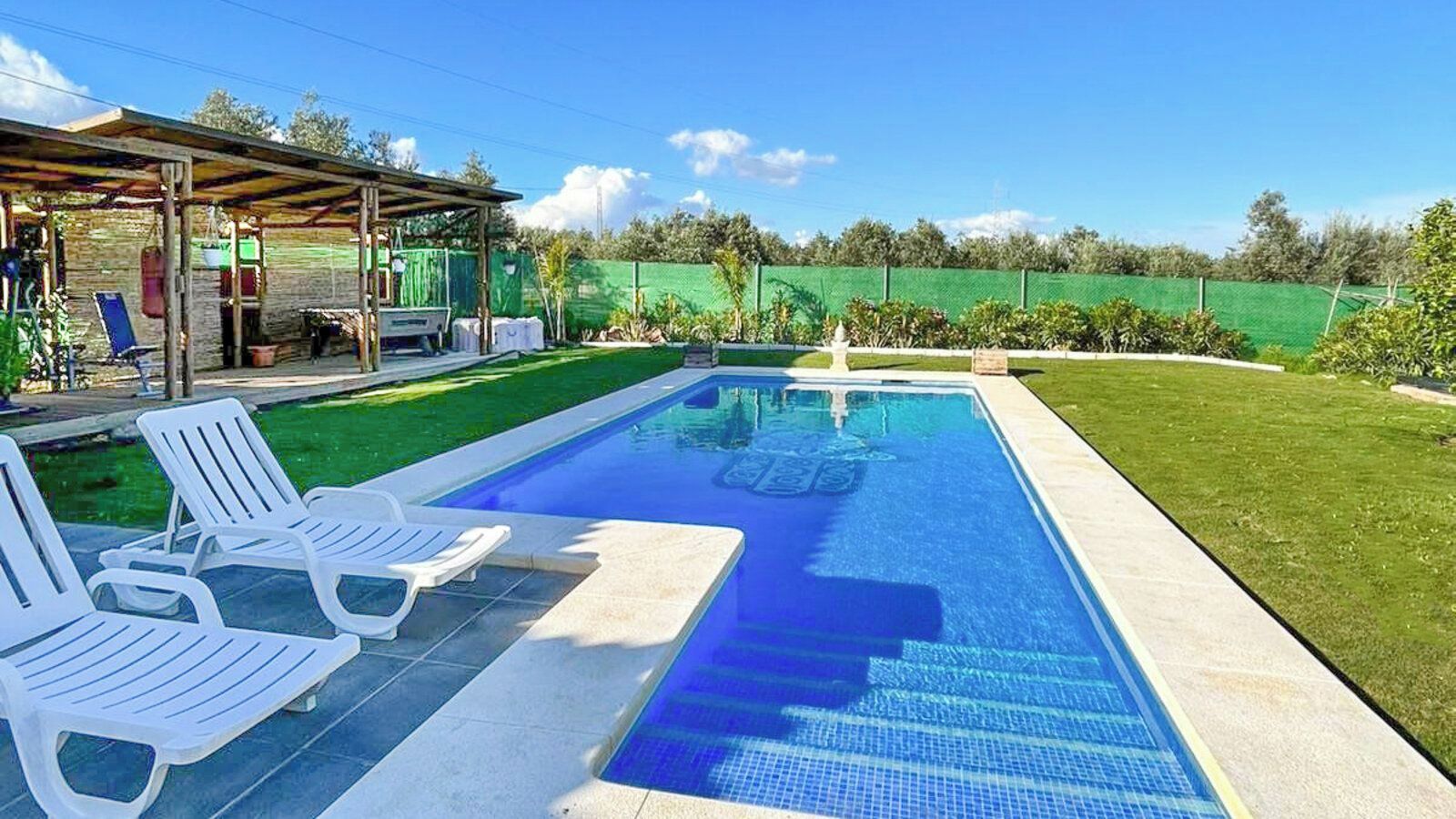A rectangular swimming pool with clear blue water, surrounded by a tiled deck. There are white lounge chairs along the poolside.