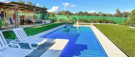 Una piscina rectangular con agua cristalina, rodeada por una solana con baldosas. Hay sillas de descanso blancas a lo largo de la piscina.