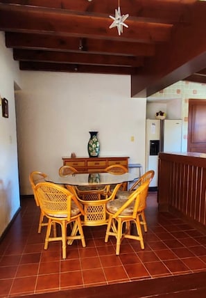 Un coin repas avec une table ronde et quatre chaises en osier, un vase posé sur la table, et une cuisine en arrière-plan.