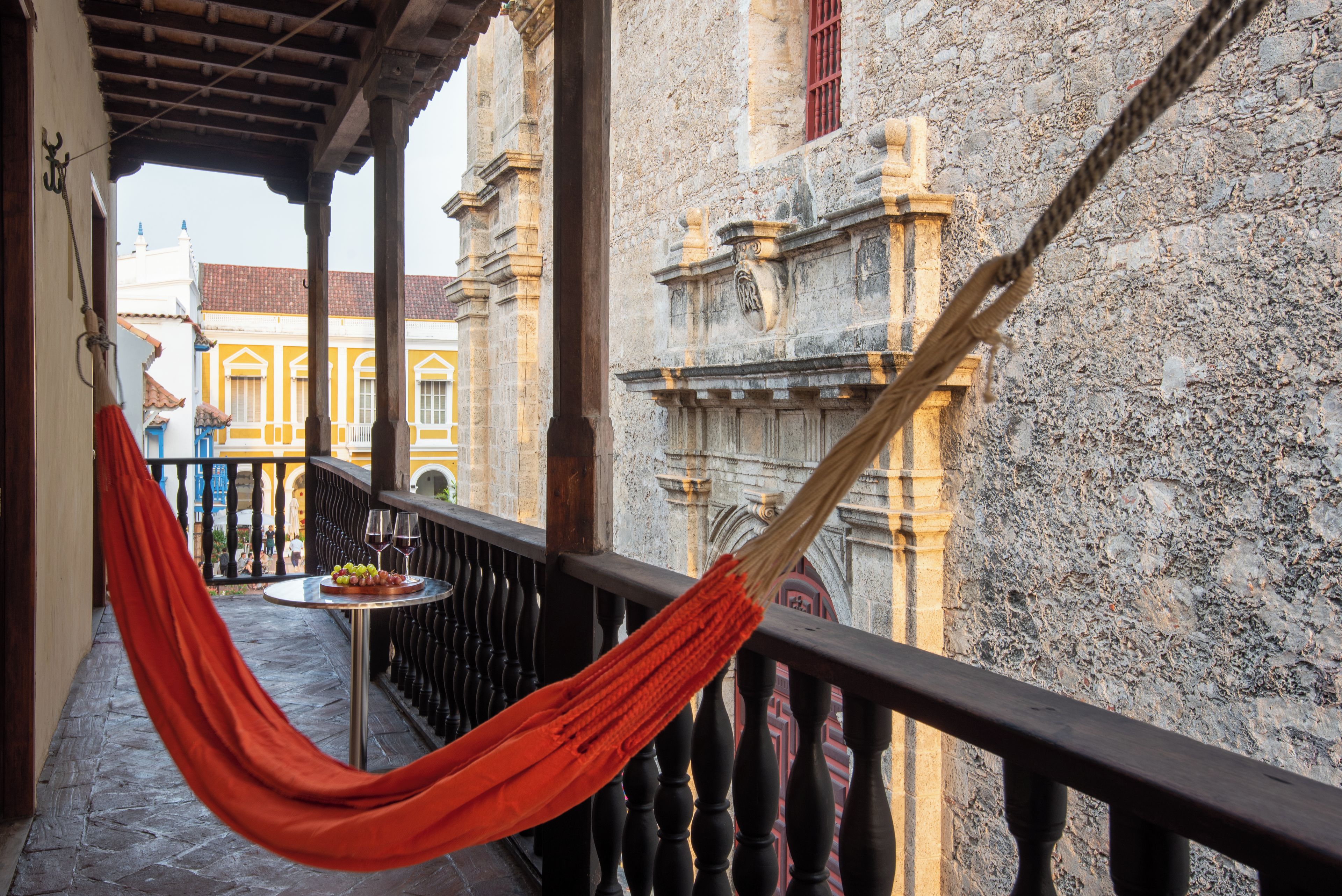 A balcony with a hammock, a small round table with a plate of grapes and two wine glasses, and a view of historic buildings.