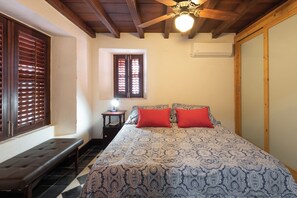 Ein Schlafzimmer mit einem Bett, zwei roten Kissen, einer Bank, einem kleinen Tisch mit einer Lampe, einem Fenster mit Jalousien und einem Deckenventilator.