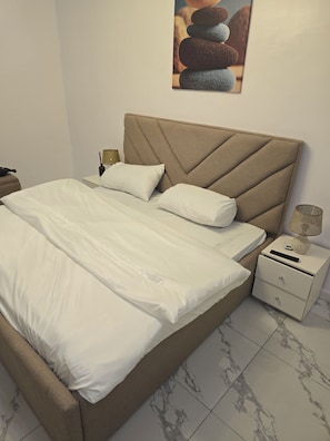 Una cama bien hecha con sábanas blancas, un cabecero con un diseño geométrico, una lámpara de noche y un cuadro en la pared de piedras apiladas.