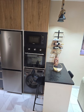 Cocina compacta con refrigerador, microondas, horno y un pequeño comedor con una silla y un cuenco decorativo.