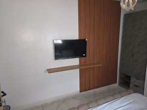 A modern bedroom with a flat-screen TV mounted on the wall, a wooden shelf below it, and a bed with a white headboard. - Royal Crowns Apartments. Cosy 2 bedroom ground floor flat in secure estate (Lagos)