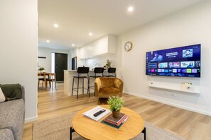 Uma sala de estar moderna com TV de tela plana, sofá, mesa de centro com um livro e controle remoto, e uma cozinha ao fundo.