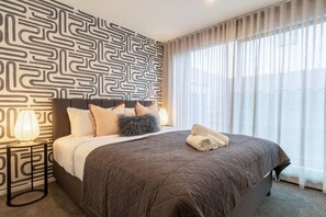 Quarto de hotel com parede estampada, cama com colcha acolchoada, duas almofadas, uma almofada fofa, abajur e janela com cortinas translúcidas.