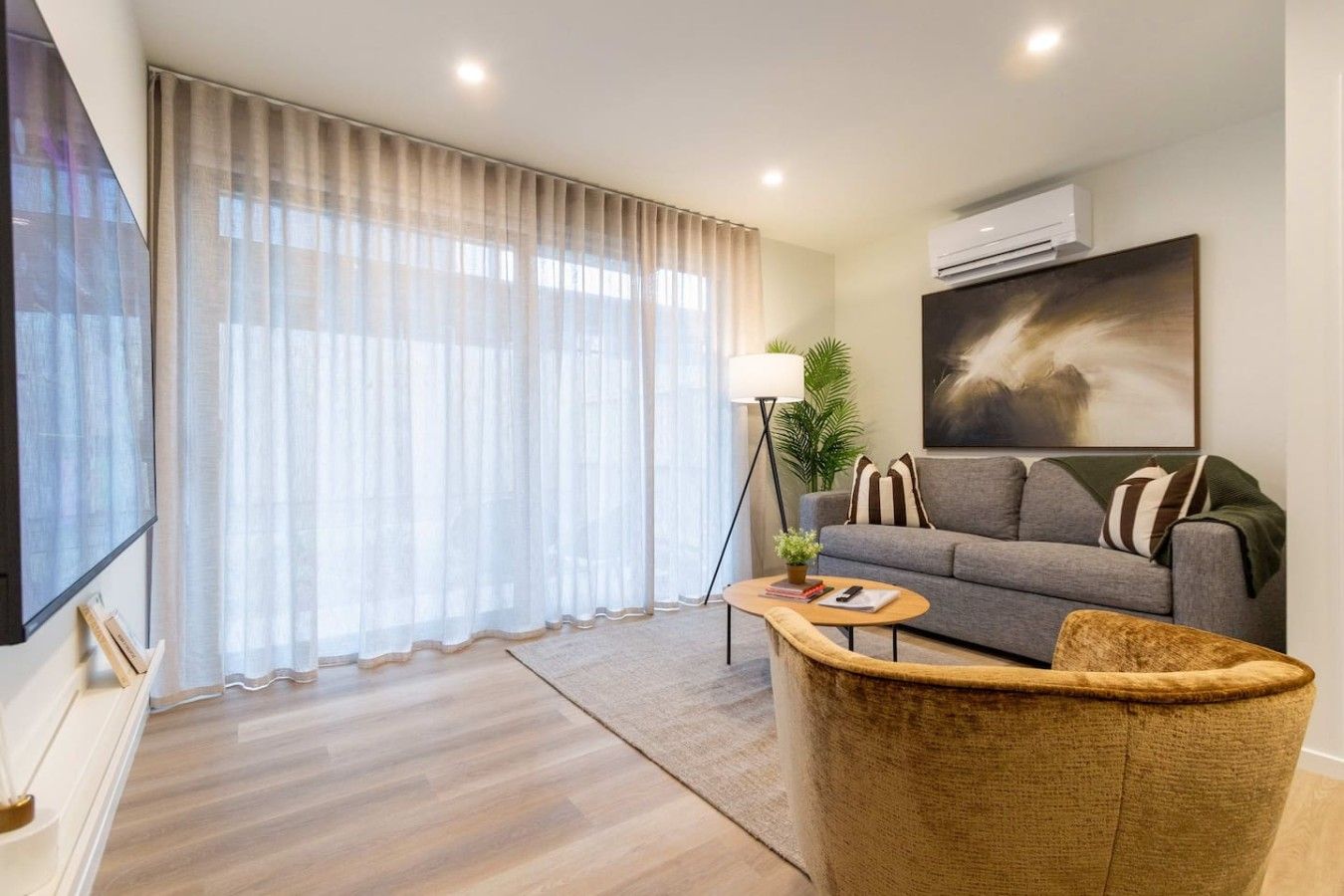 Una sala moderna con un sofá gris, una mesa de centro, una lámpara de pie y un ventanal con cortinas translúcidas.