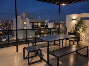 Una terraza en la azotea con mesa, bancos, una planta en maceta y vistas a la ciudad al anochecer.