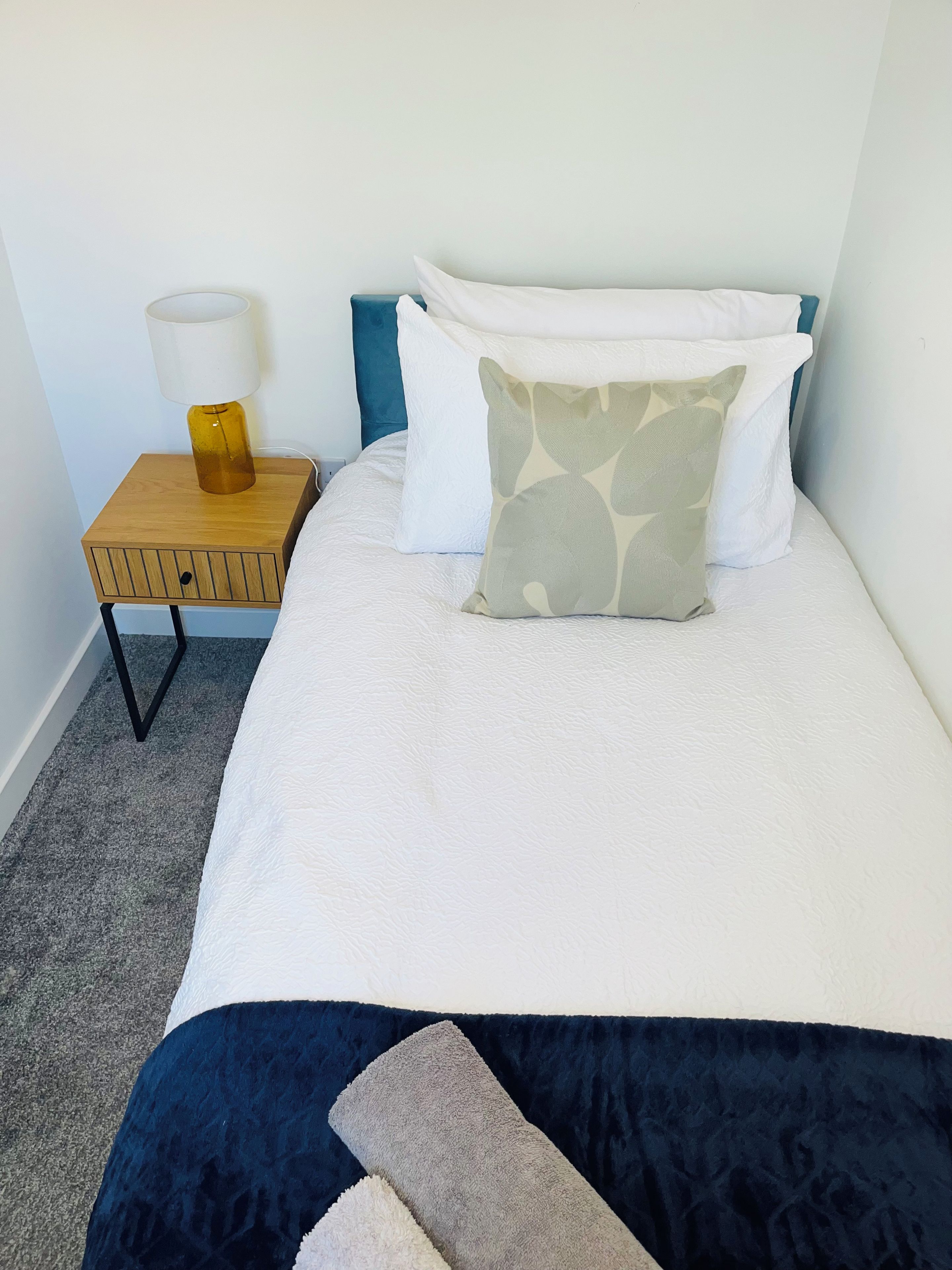 Een net opgemaakt bed met wit beddengoed en een blauw hoofdeinde, een klein houten nachtkastje met een lamp, en een grijs vloerkleed.