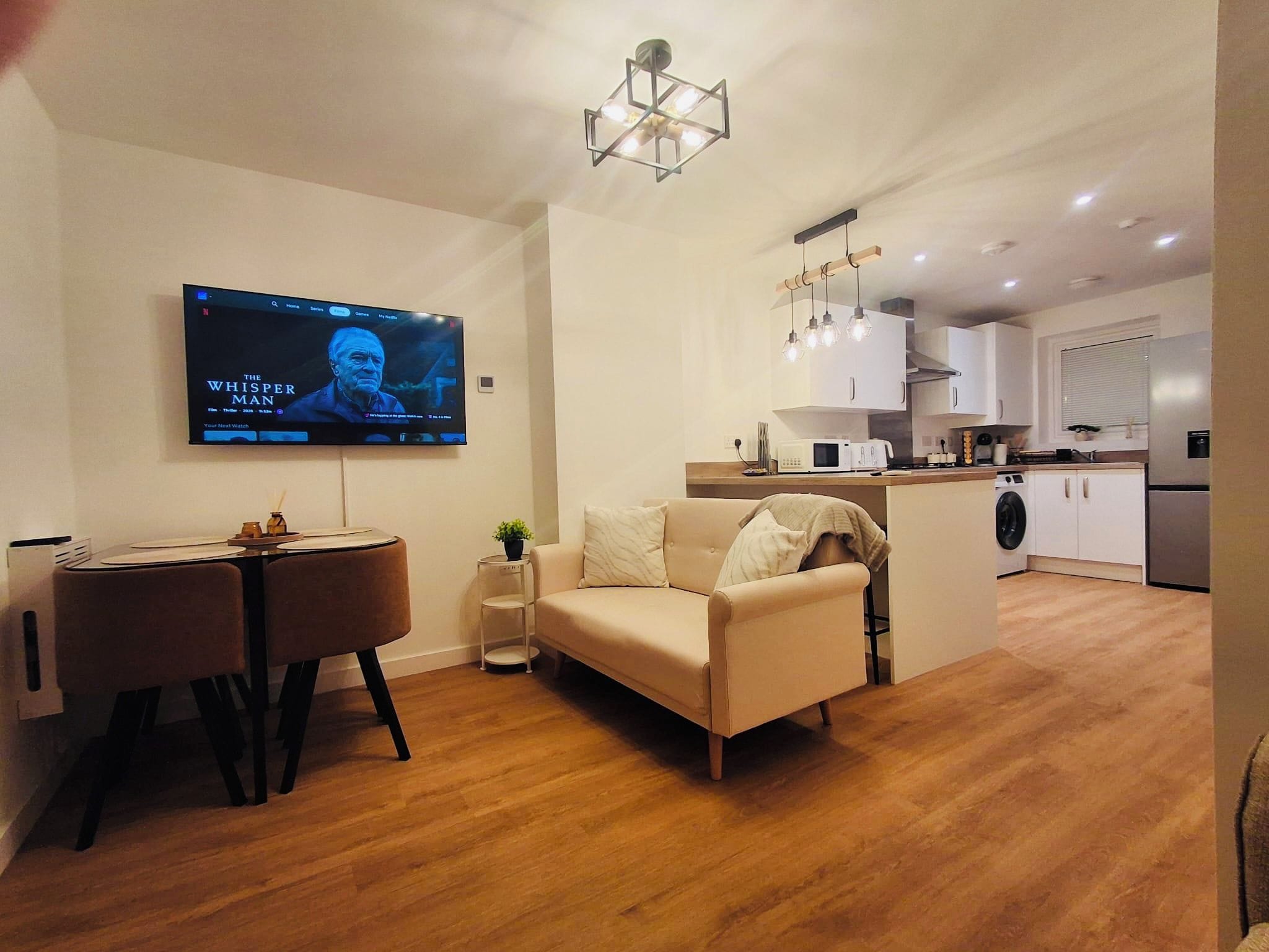 Een moderne woonkamer met een flatscreen televisie, een bank, een eettafel en een keuken met witte kasten en een magnetron.