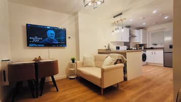 Een moderne woonkamer met een flatscreen televisie, een bank, een eettafel en een keuken met witte kasten en een magnetron.