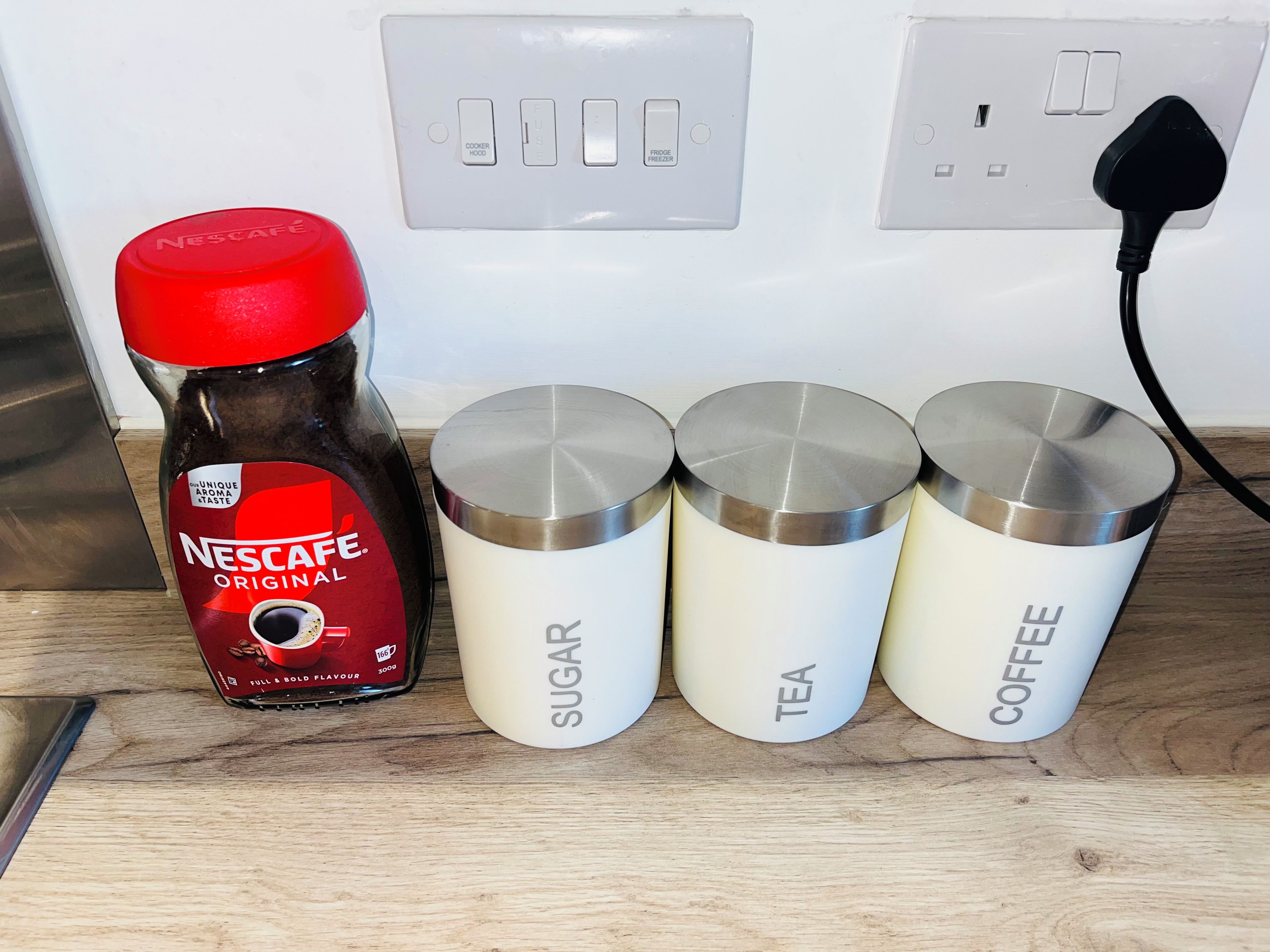 Uma bancada de cozinha com um pote de café Nescafé Original, açúcar, chá e recipientes para café.