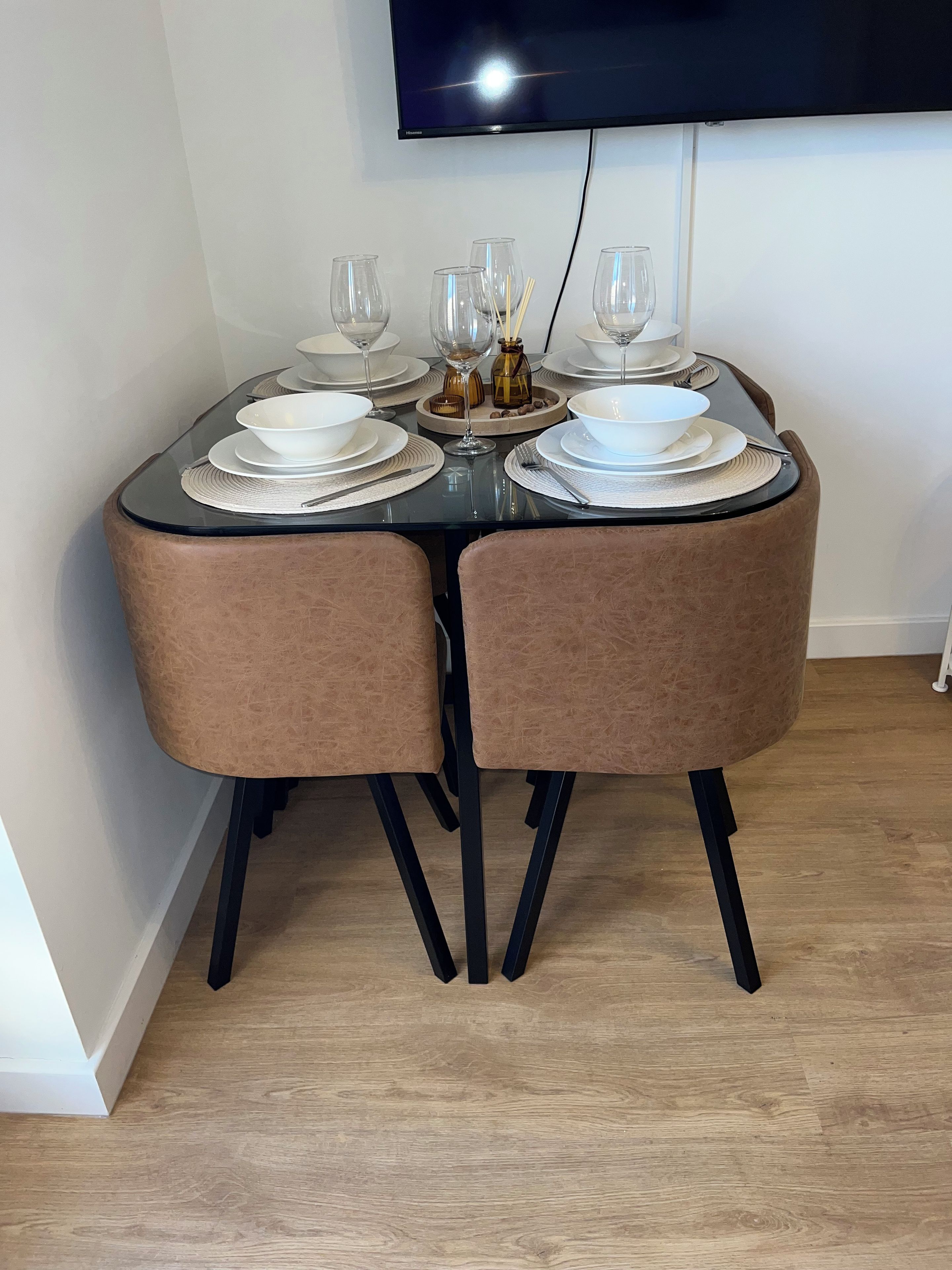 Een eettafel voor vier personen, gedekt met witte borden, kommen en bestek, inclusief wijnglazen en een klein decoratief object.
