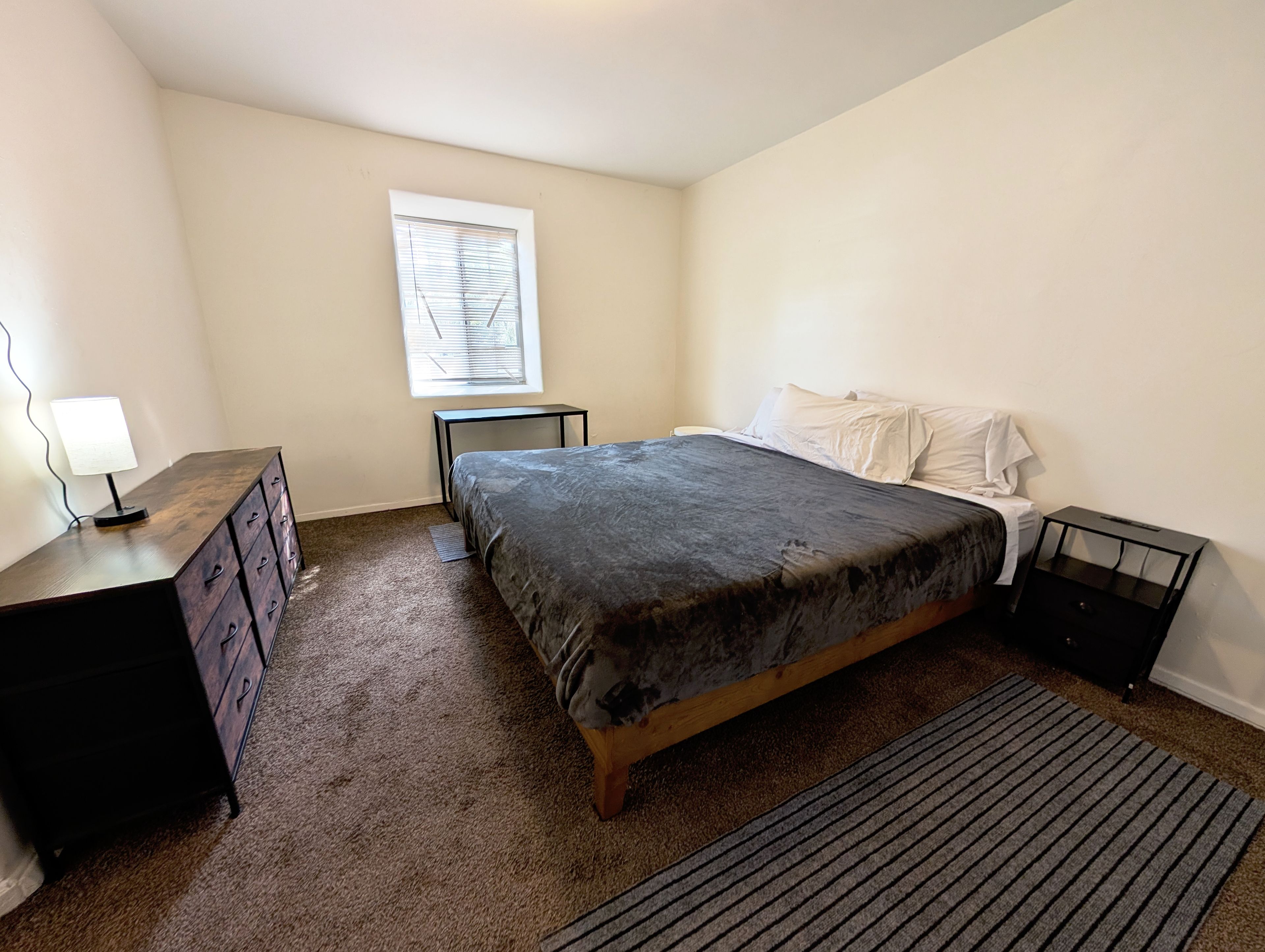 A bedroom with a bed, a dresser, a nightstand, a lamp, and a window.