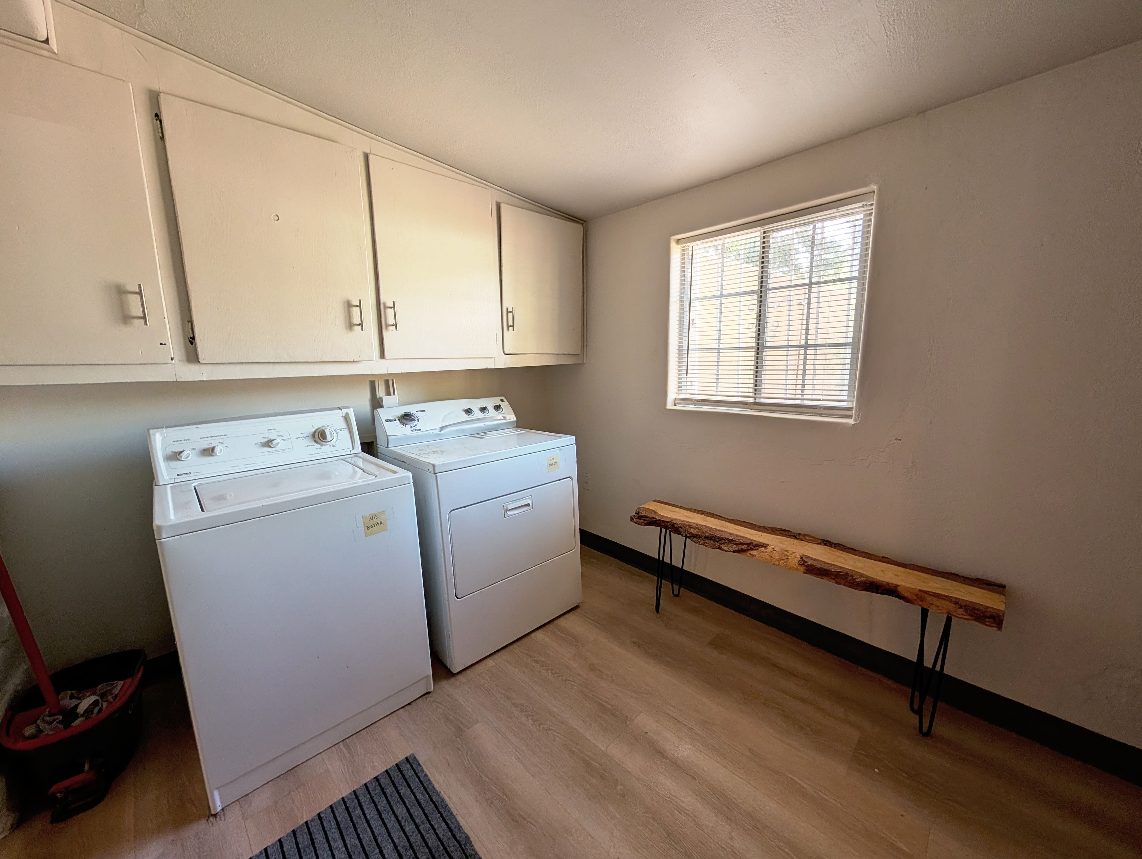 A laundry room with two white washers and dryers, wooden cabinets, a window with blinds, and a bench.